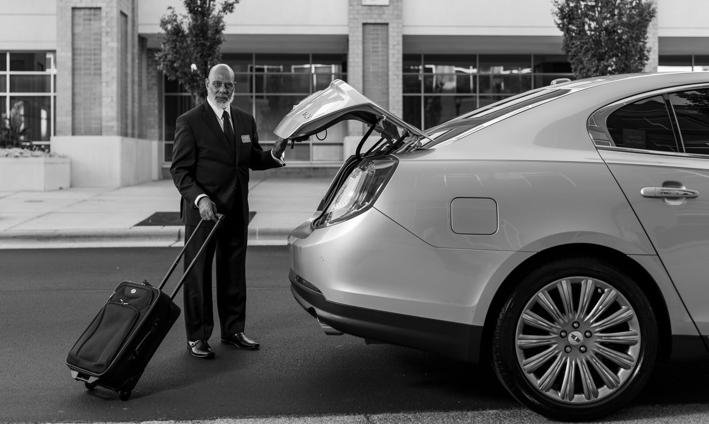 A chauffeur in a suit loading a suitcase into the trunk of a sedan outside an airport terminal