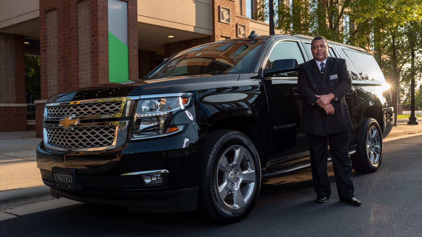 A uniformed chauffeur standing beside a black full-size SUV outside a building at golden hour