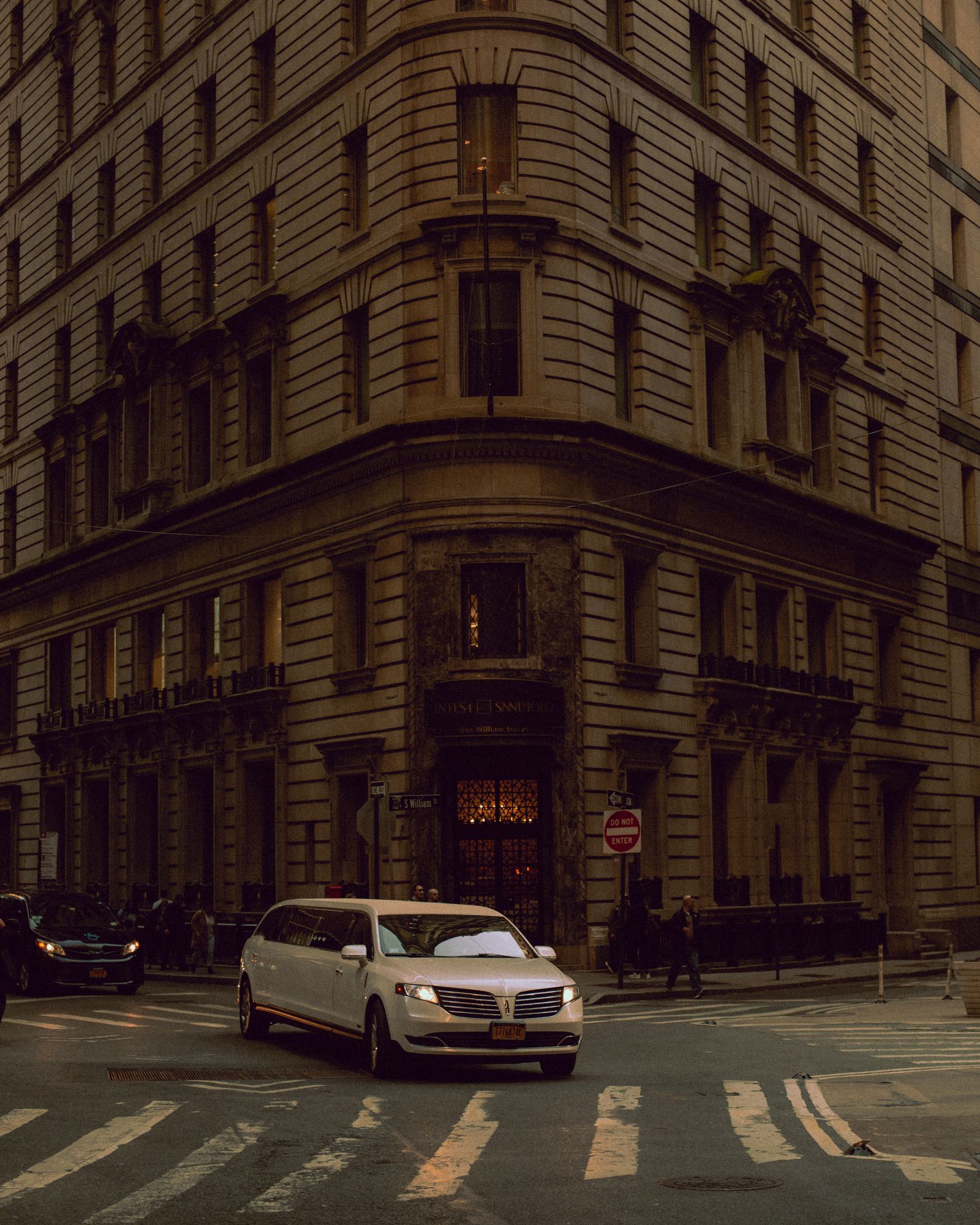 A white stretch limousine crossing an empty downtown intersection between stone office towers at dusk