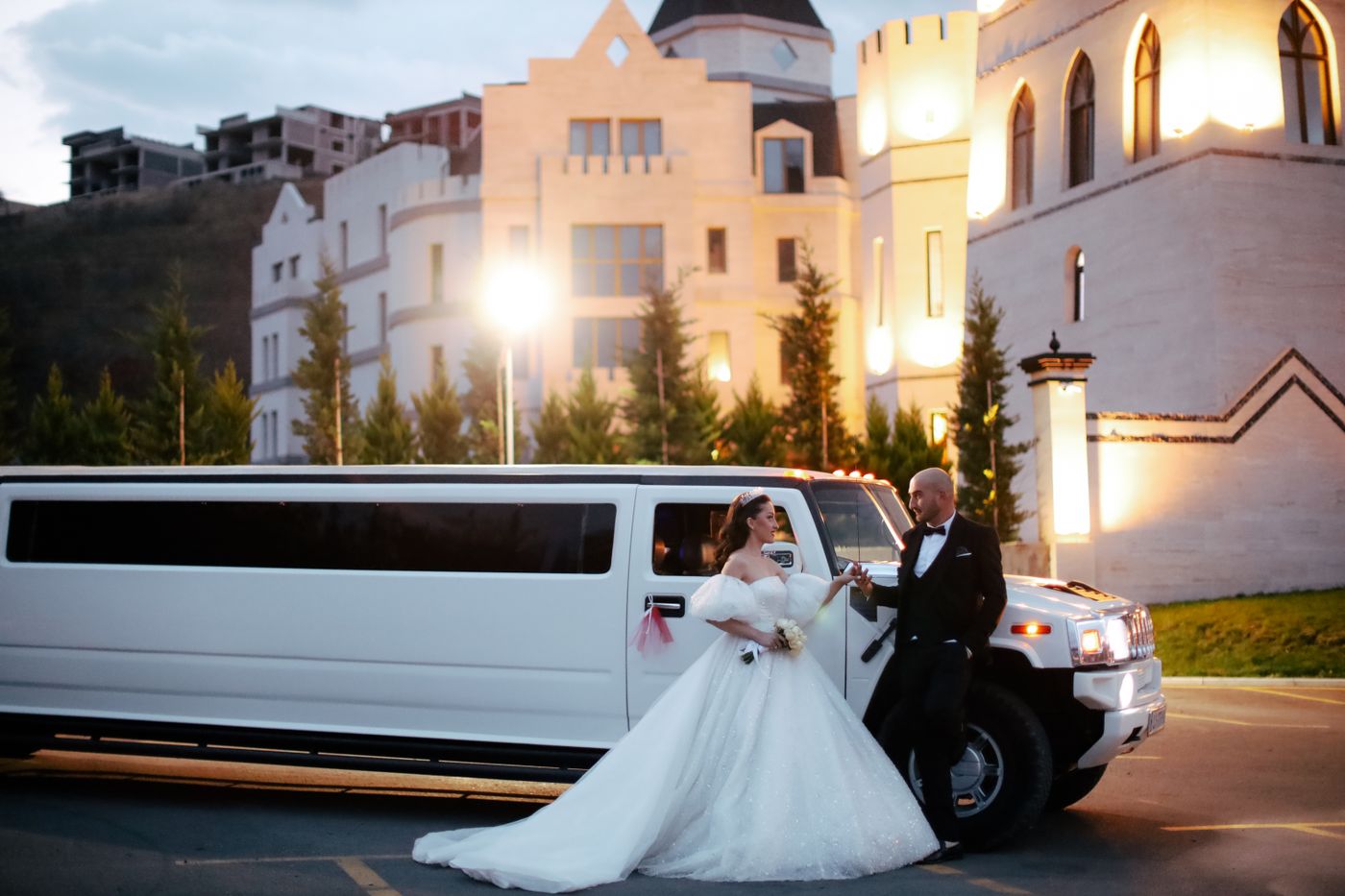 A bride and groom standing beside a white SUV stretch limousine outside an illuminated venue at dusk